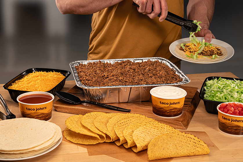 Person assembling tacos from a catering spread with seasoned ground beef, shredded cheese, lettuce, tortillas, taco shells, and sides from Taco John’s on a wooden table.